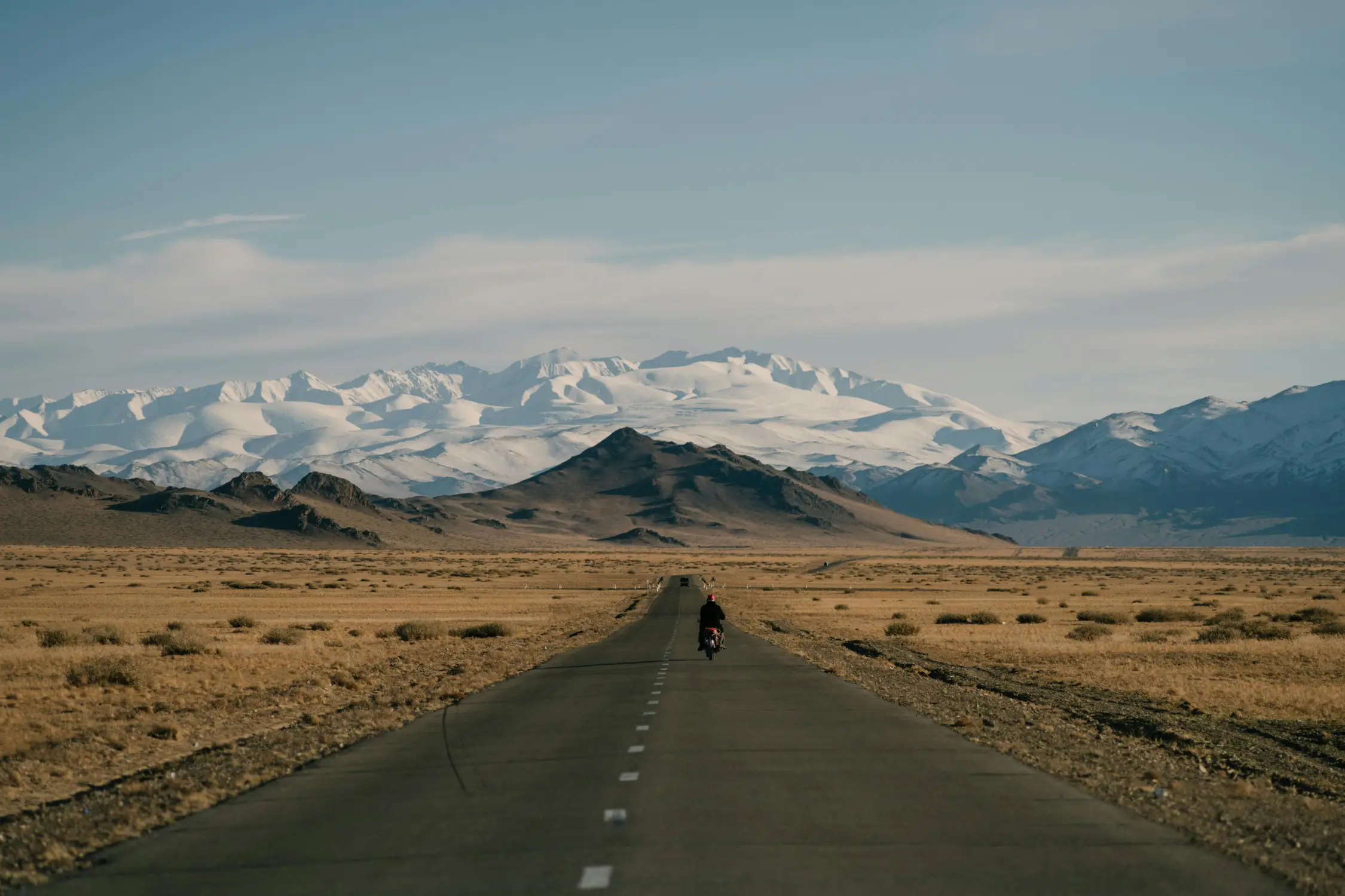 Motorcyclist riding alone on a straight empty road through the Mongolian steppe toward snow-capped Altai mountains under a wide blue sky