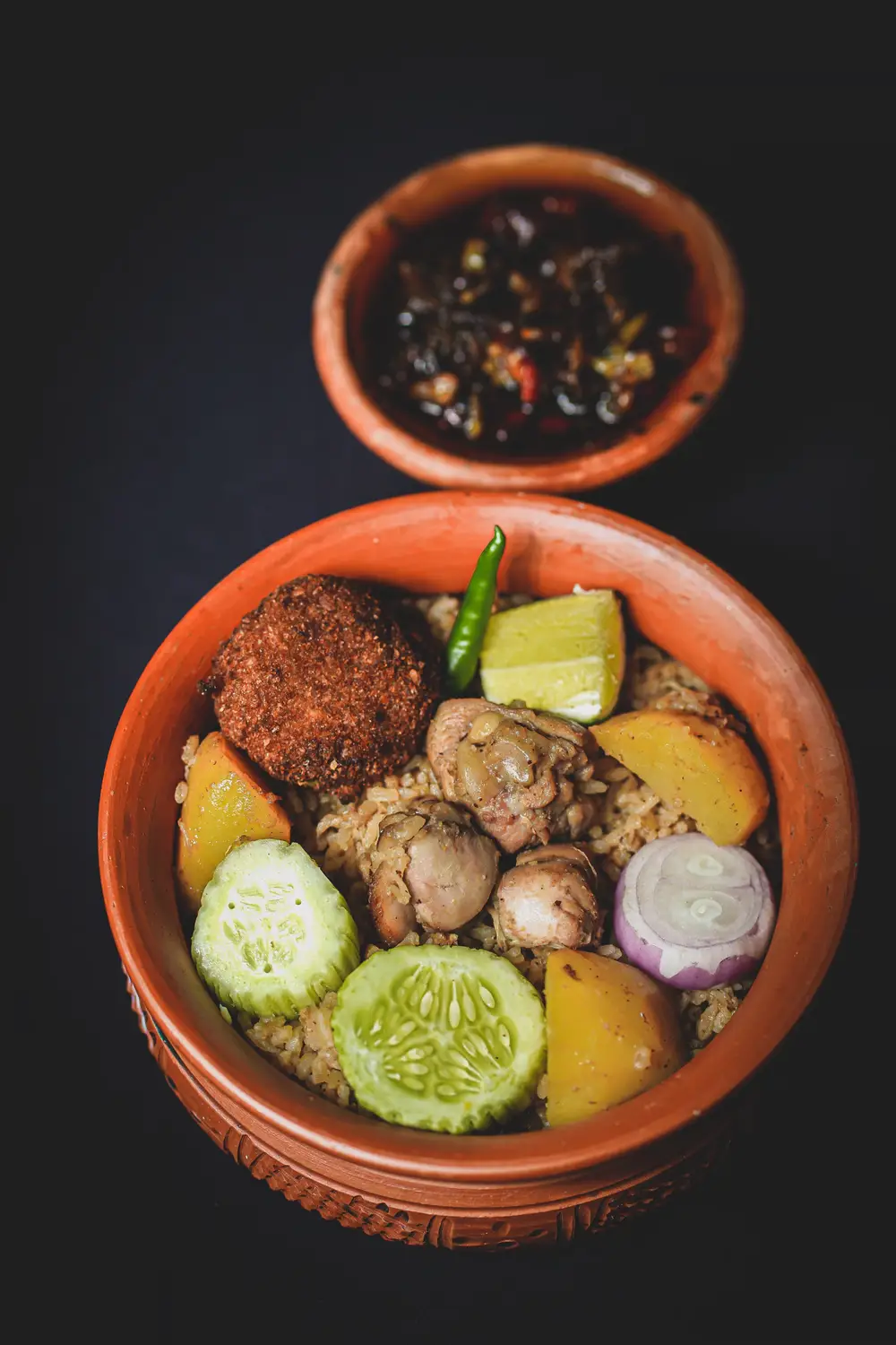 Traditional Bangladeshi Kacchi Biryani served in a clay pot with boiled egg, potato, cucumber, onion, green chili, and dark tamarind chutney on the side