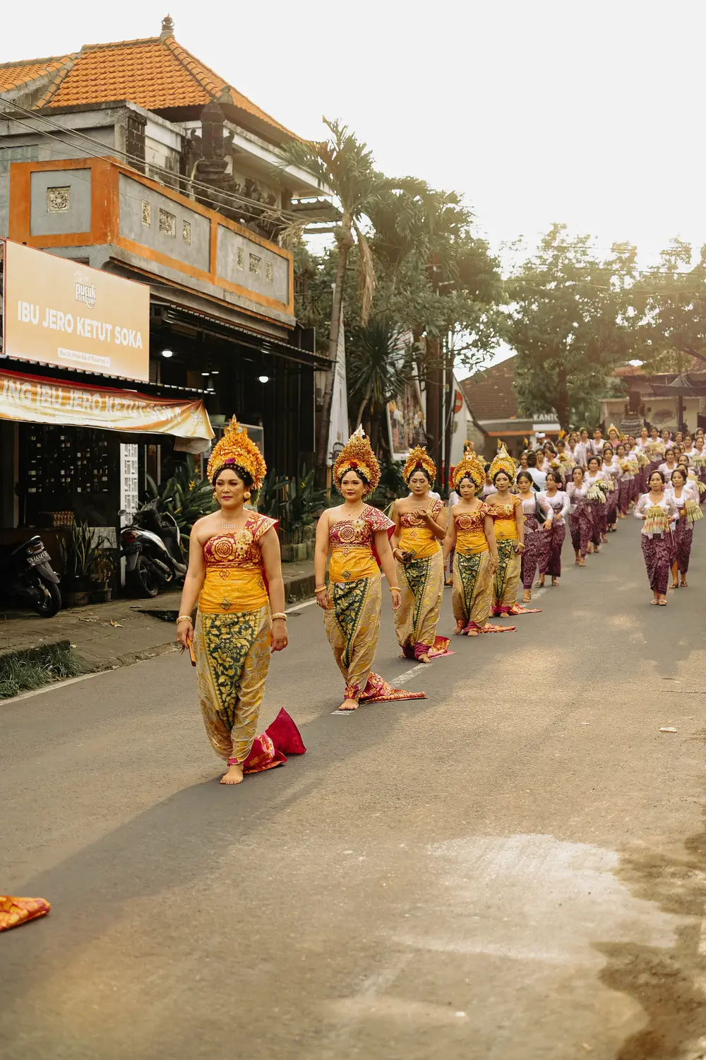 Balinese women in traditional golden dress and headdresses walking in a ceremonial procession along a village street in Bali, Indonesia.