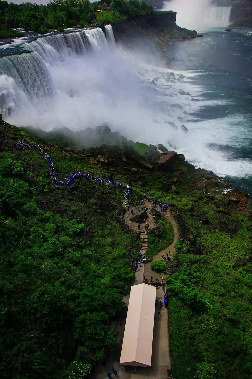 Aerial view of Niagara Falls with mist rising from the cascading water, and a walkway winding through greenery toward the falls.