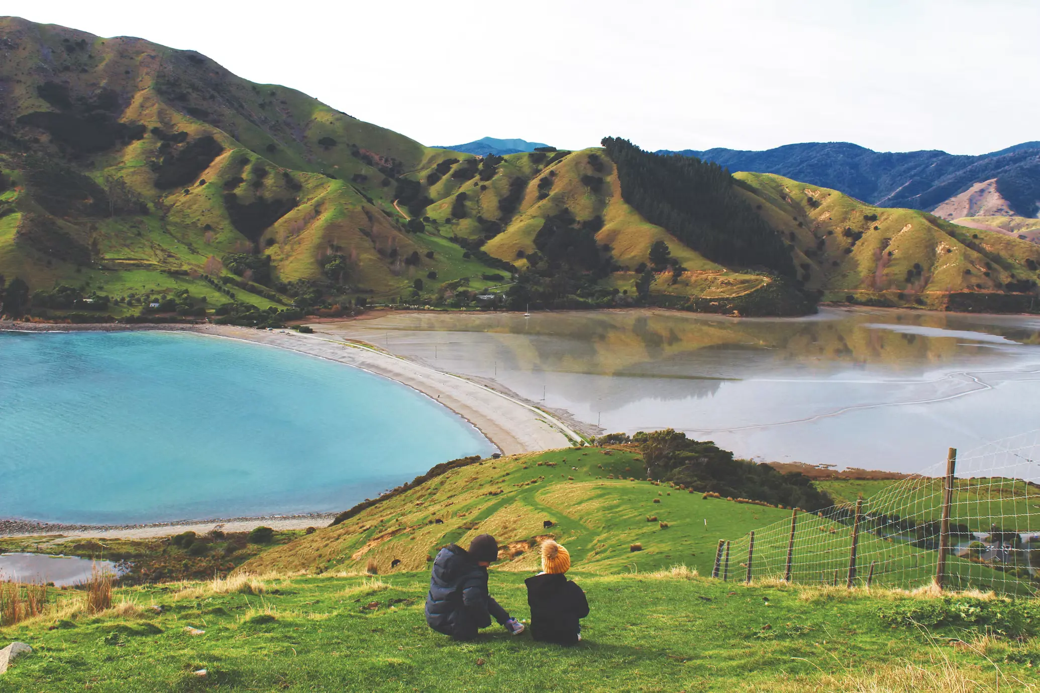 Two people sitting on a grassy hillside overlooking a curved sandy bay, turquoise water, and rolling green hills.