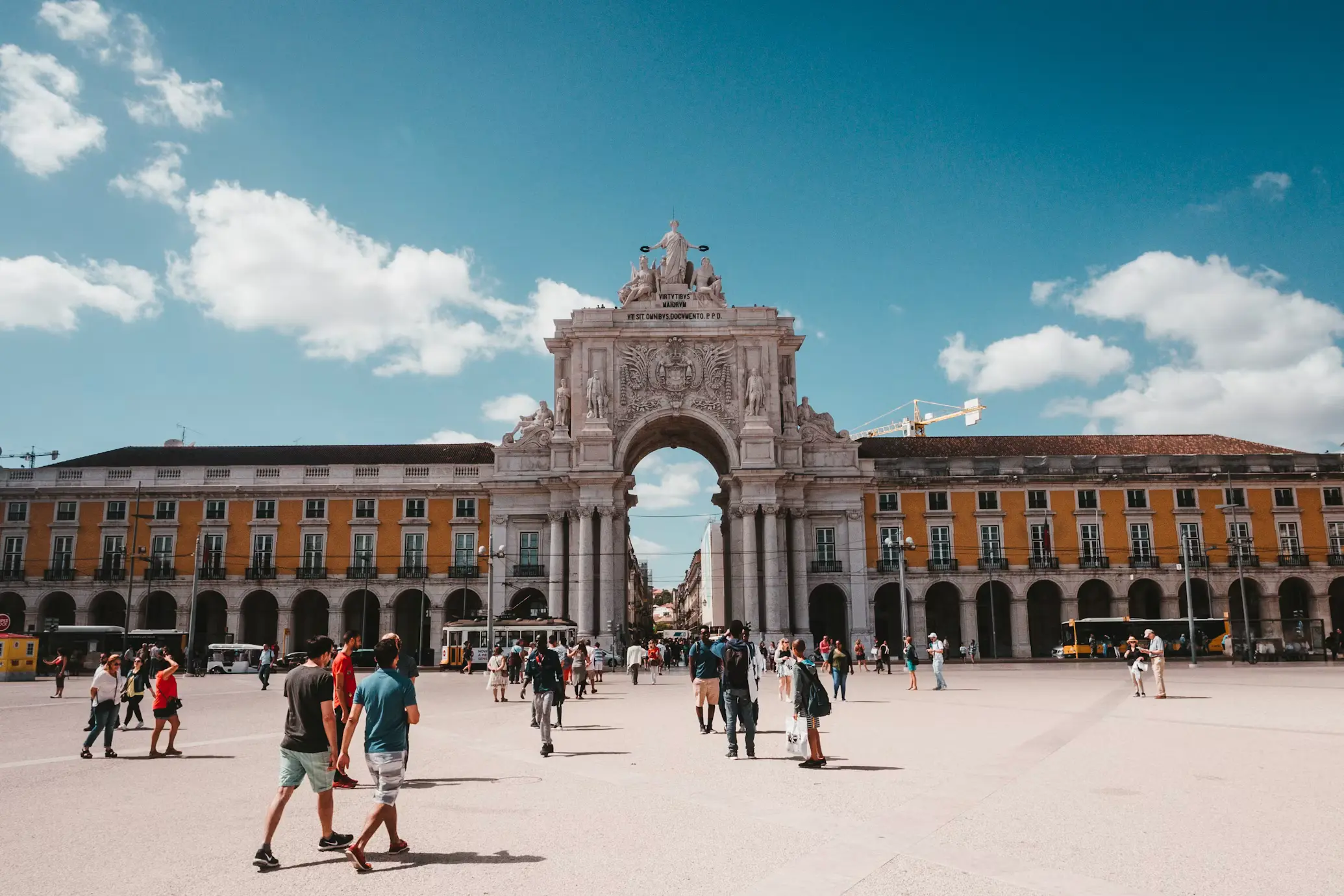 Grand stone triumphal arch in a large city square, with people walking beneath it under a bright blue sky.