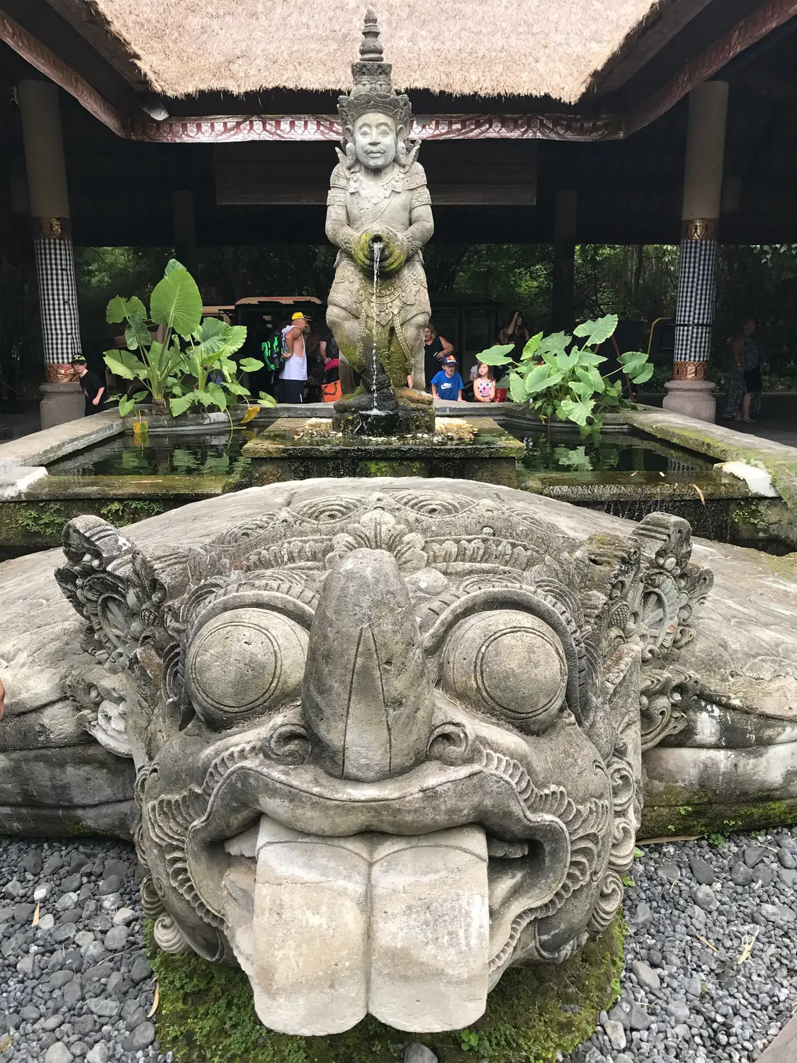 Ornate Balinese stone fountain statue with a fierce carved face and protruding tongue, set in a temple courtyard garden in Bali, Indonesia.
