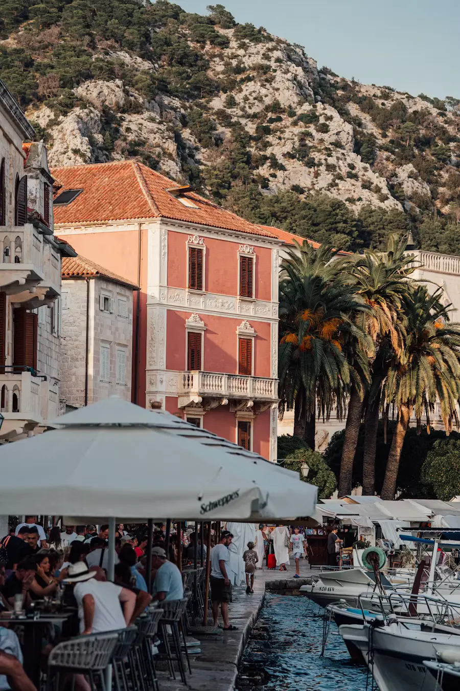 Outdoor waterfront café with people dining under umbrellas, colorful buildings and palm trees along the harbor.