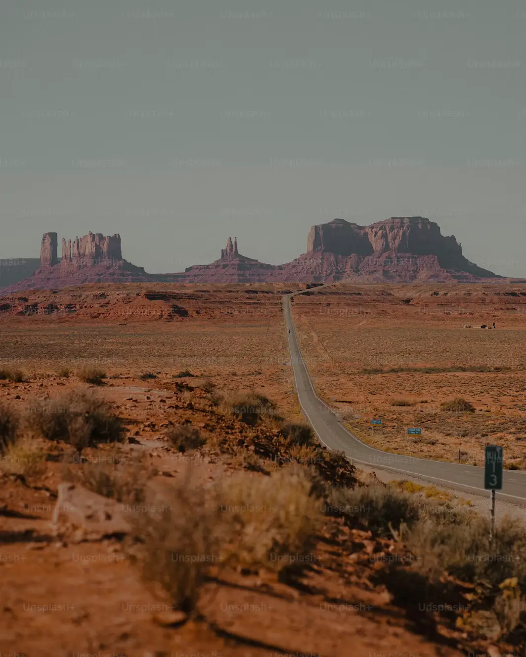 A long straight desert highway stretching toward the sandstone buttes of Monument Valley on a hazy day.