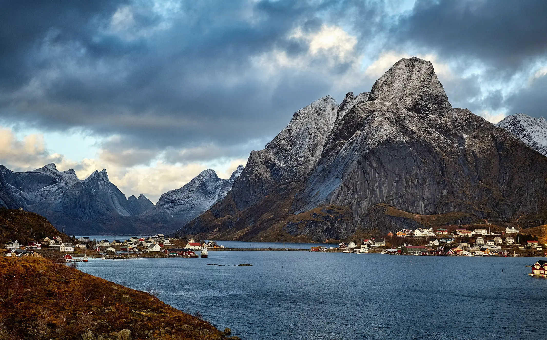 Dramatic storm clouds gathering over jagged, snow-dusted peaks above a village on the shore of a Norwegian fjord.