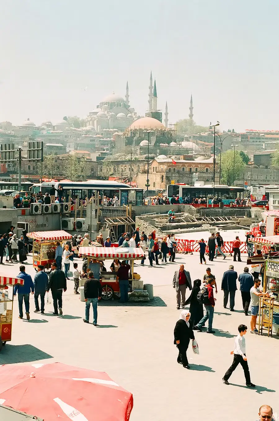 An elevated view of a busy outdoor market square in Istanbul, with mosque domes and minarets visible on the skyline in the distance.