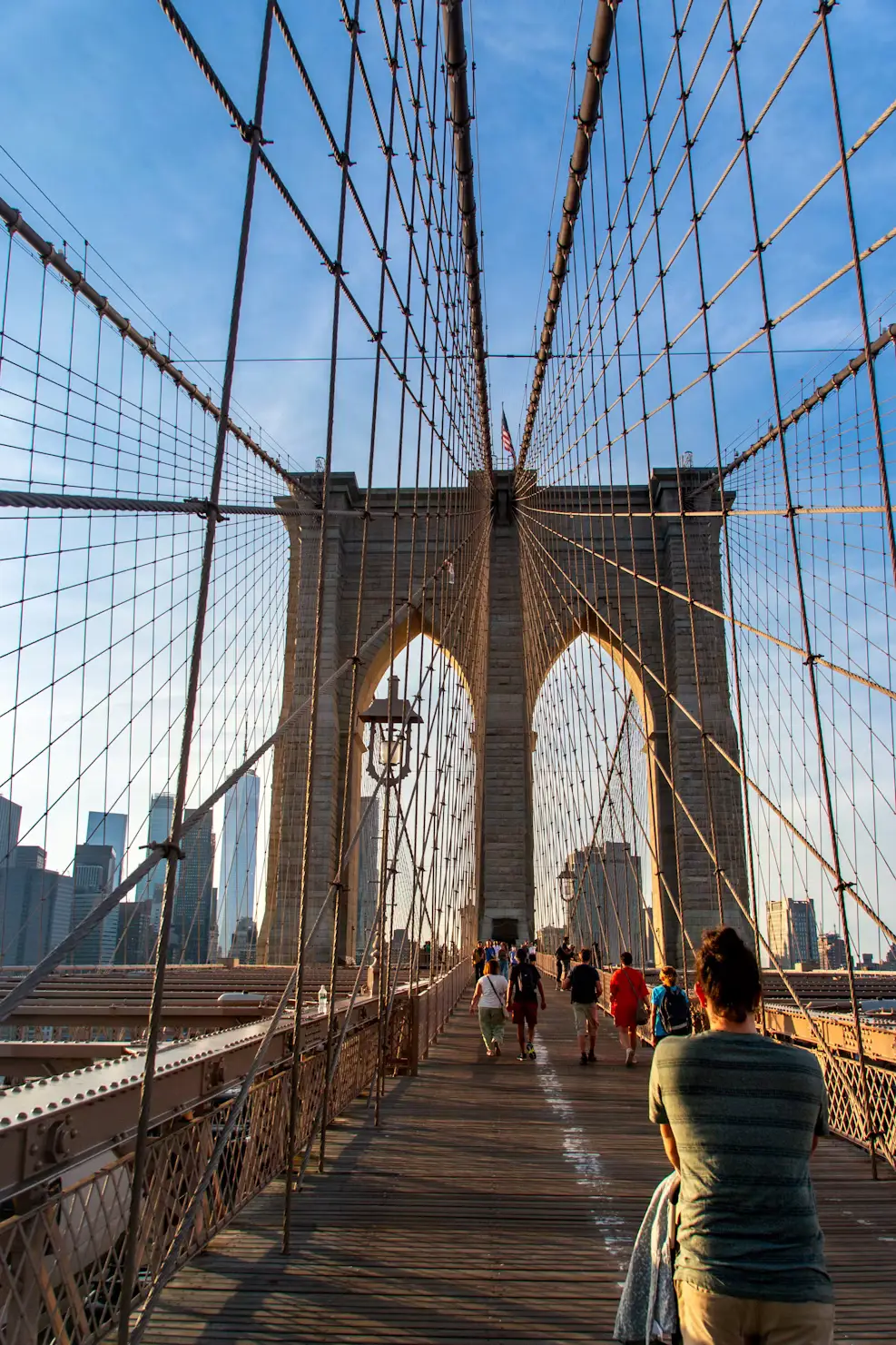 Pedestrians walking the Brooklyn Bridge's wooden promenade beneath its web of steel suspension cables.
