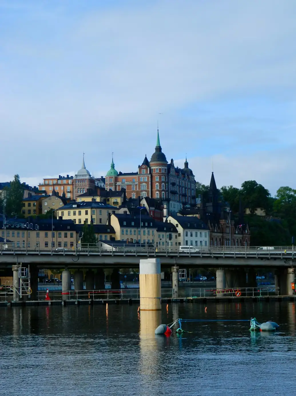 A waterfront cityscape with historic buildings and church spires along the shore, seen across calm water with a bridge pillar in the foreground.