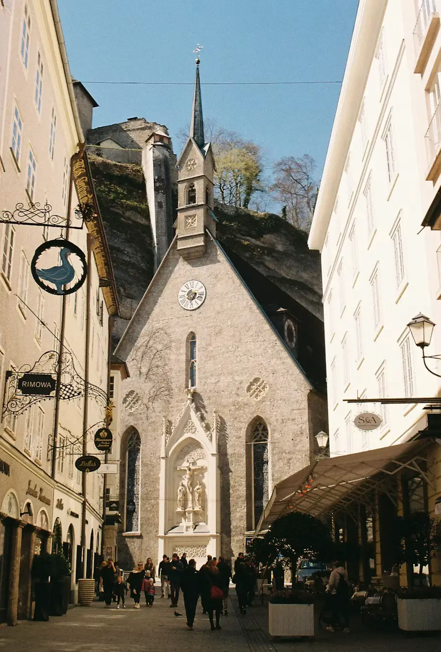 A narrow pedestrian street lined with shops, leading to a Gothic church with a tall spire, with people walking along the storefronts.