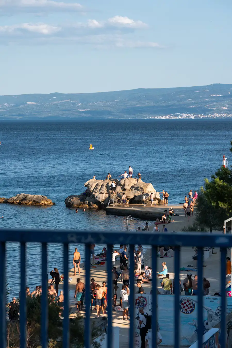People relaxing along a seaside railing with rocky cliffs and the sea in the background under a blue sky