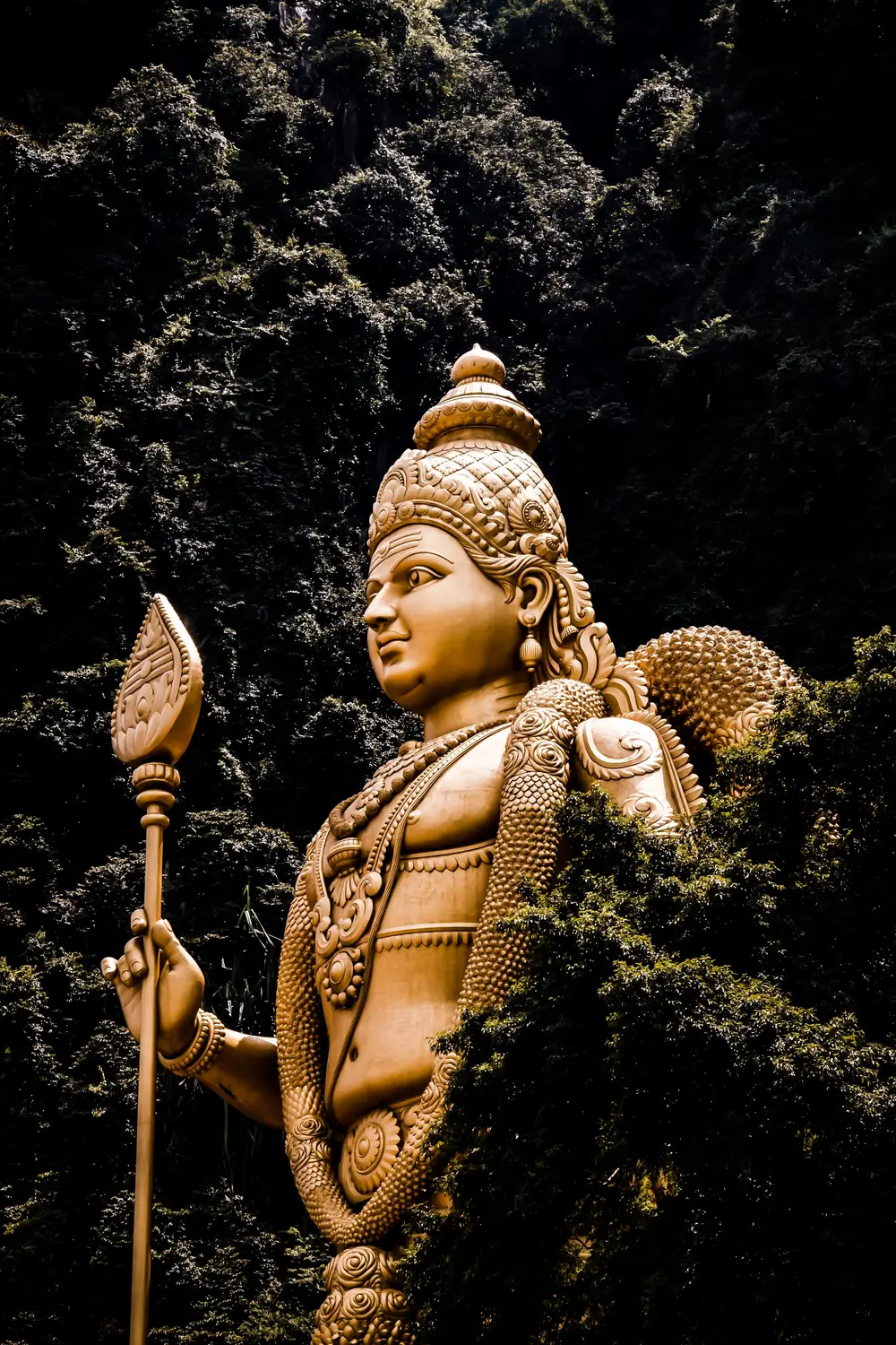 Golden bronze Lord Murugan Hindu deity statue holding vel spear against dark background at Batu Caves Malaysia