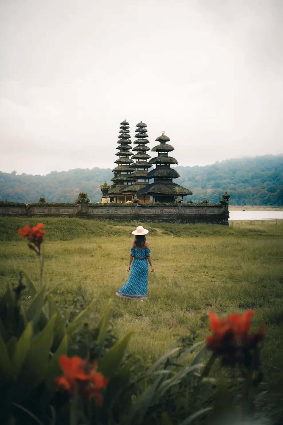 Woman in a blue dress and hat standing in a flower-filled meadow before a tiered meru temple at dawn in Bali, Indonesia.
