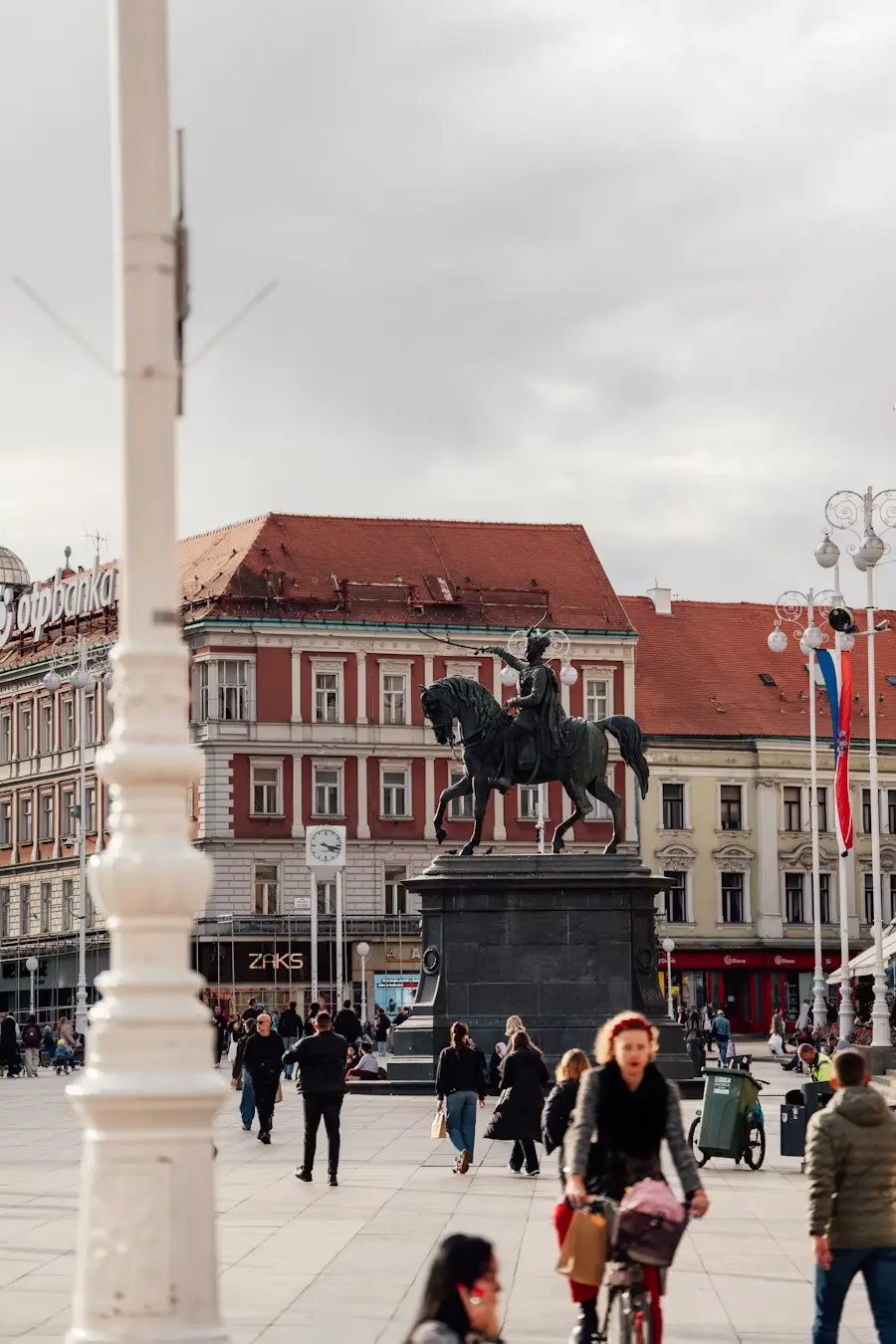 Equestrian statue in a European town square, with people walking past historic red-roofed buildings under an overcast sky.