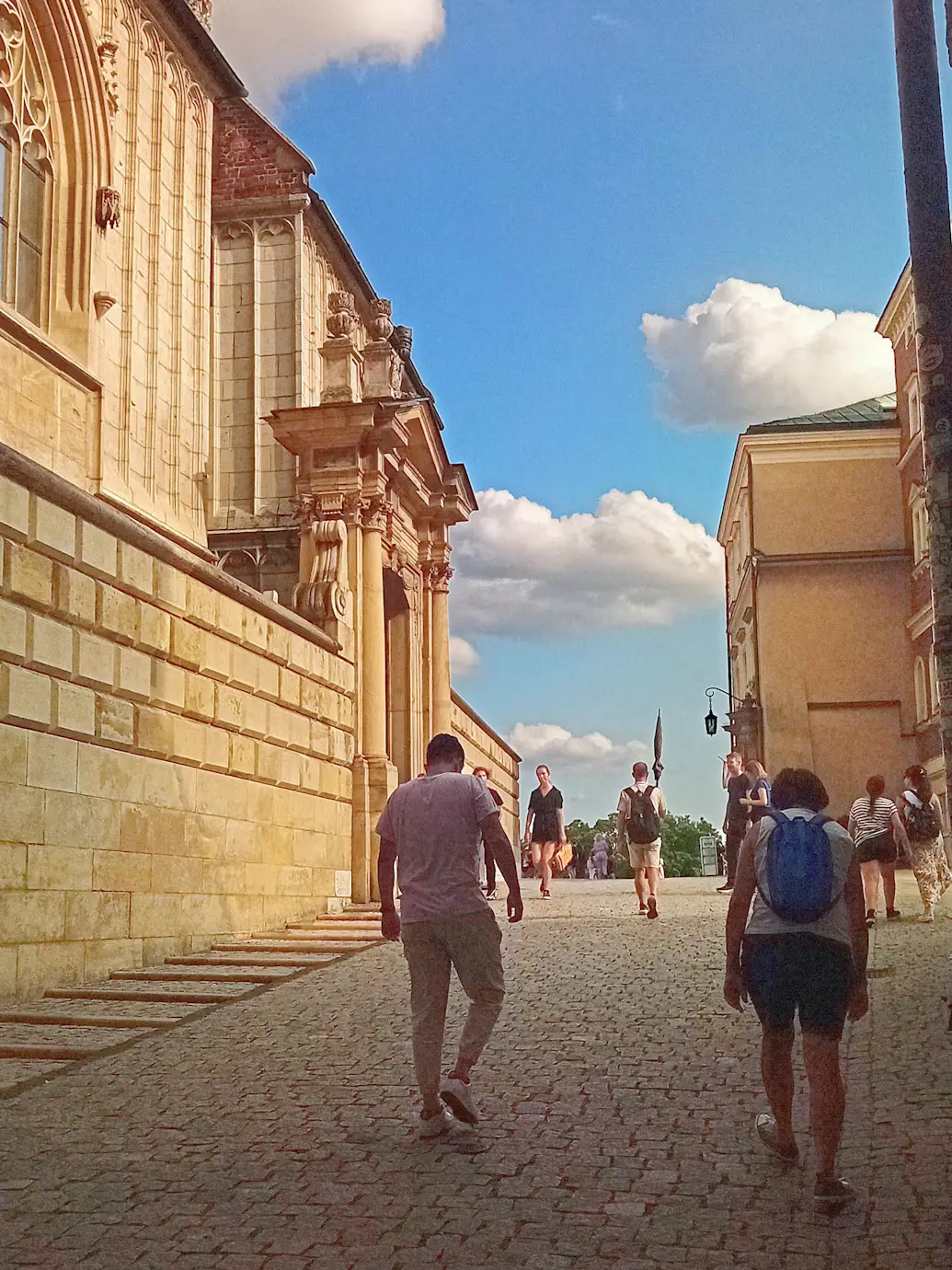 Pedestrians walking along a grand baroque building facade under a partly cloudy sky, Poland.