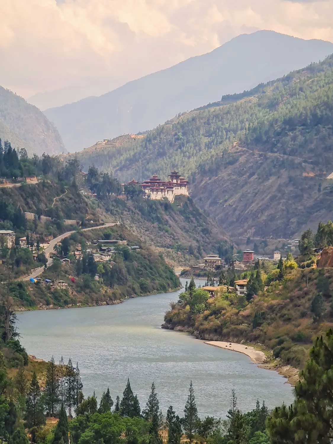 Mountain valley with a calm river, forested slopes, and a hillside dzong beneath misty peaks in Bhutan.