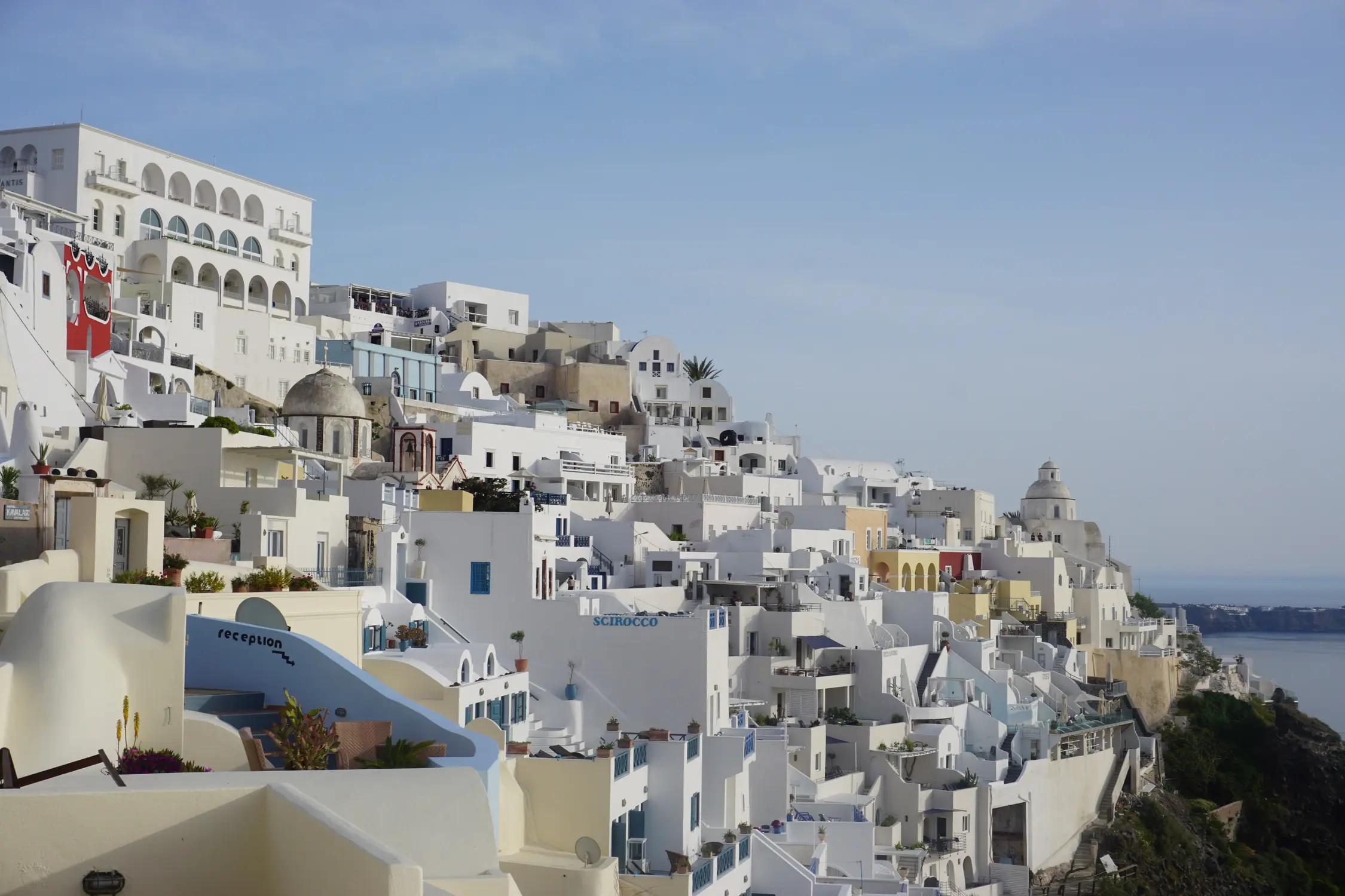 Whitewashed hillside village with domed buildings and terraces overlooking the sea under a hazy blue sky.