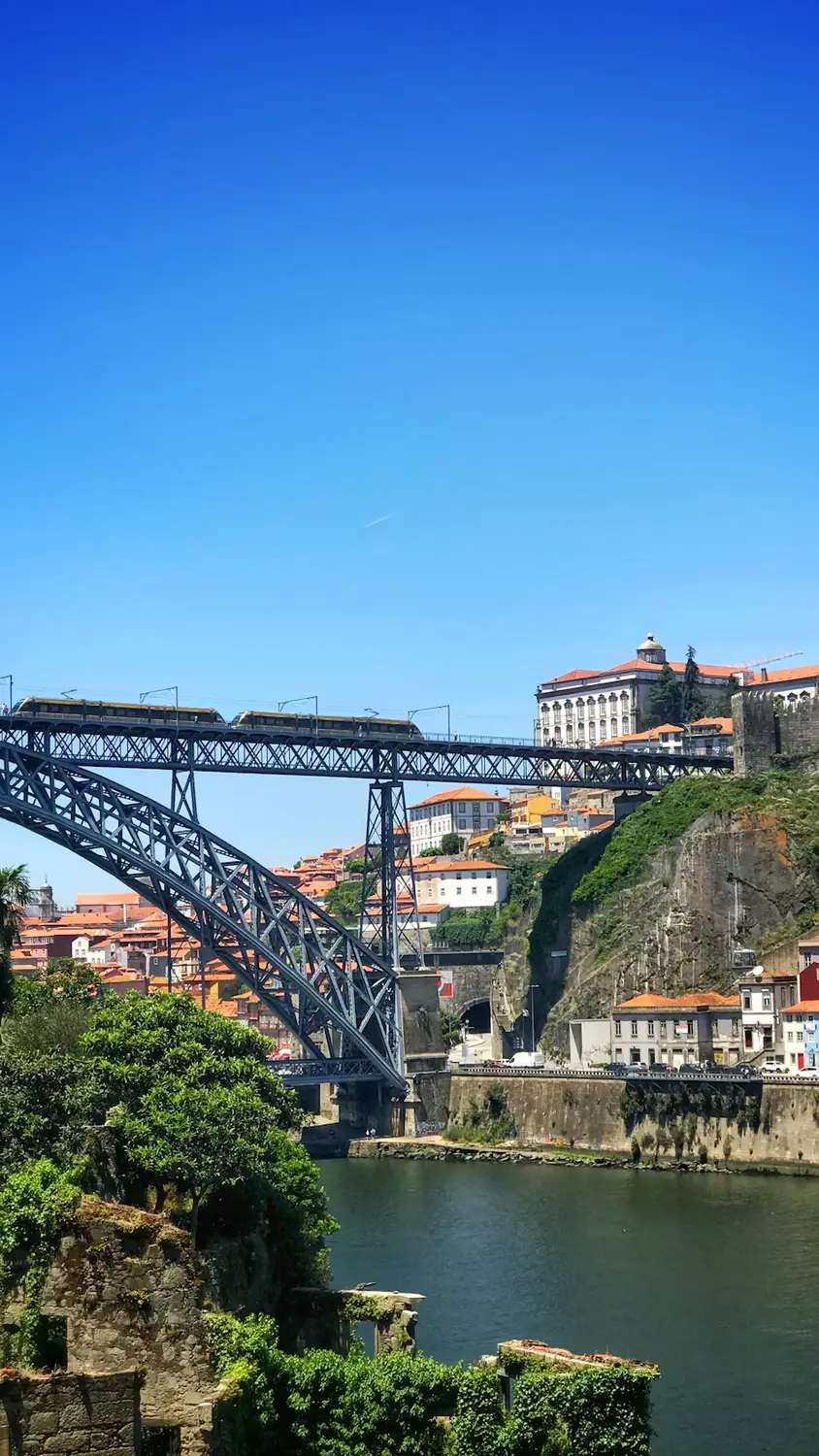 Steel arch bridge spanning a river, with a train crossing above and a hillside city of terracotta-roofed buildings in the background.