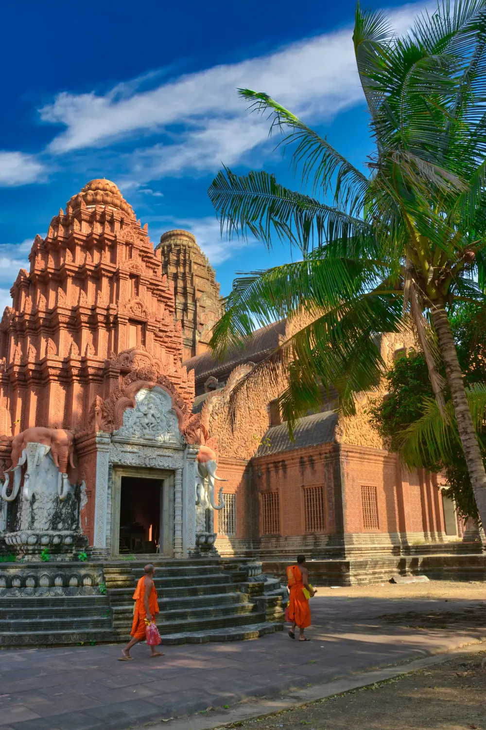 Two Buddhist monks in orange robes walking past ornate red sandstone temple entrance with white elephant statues, Cambodia