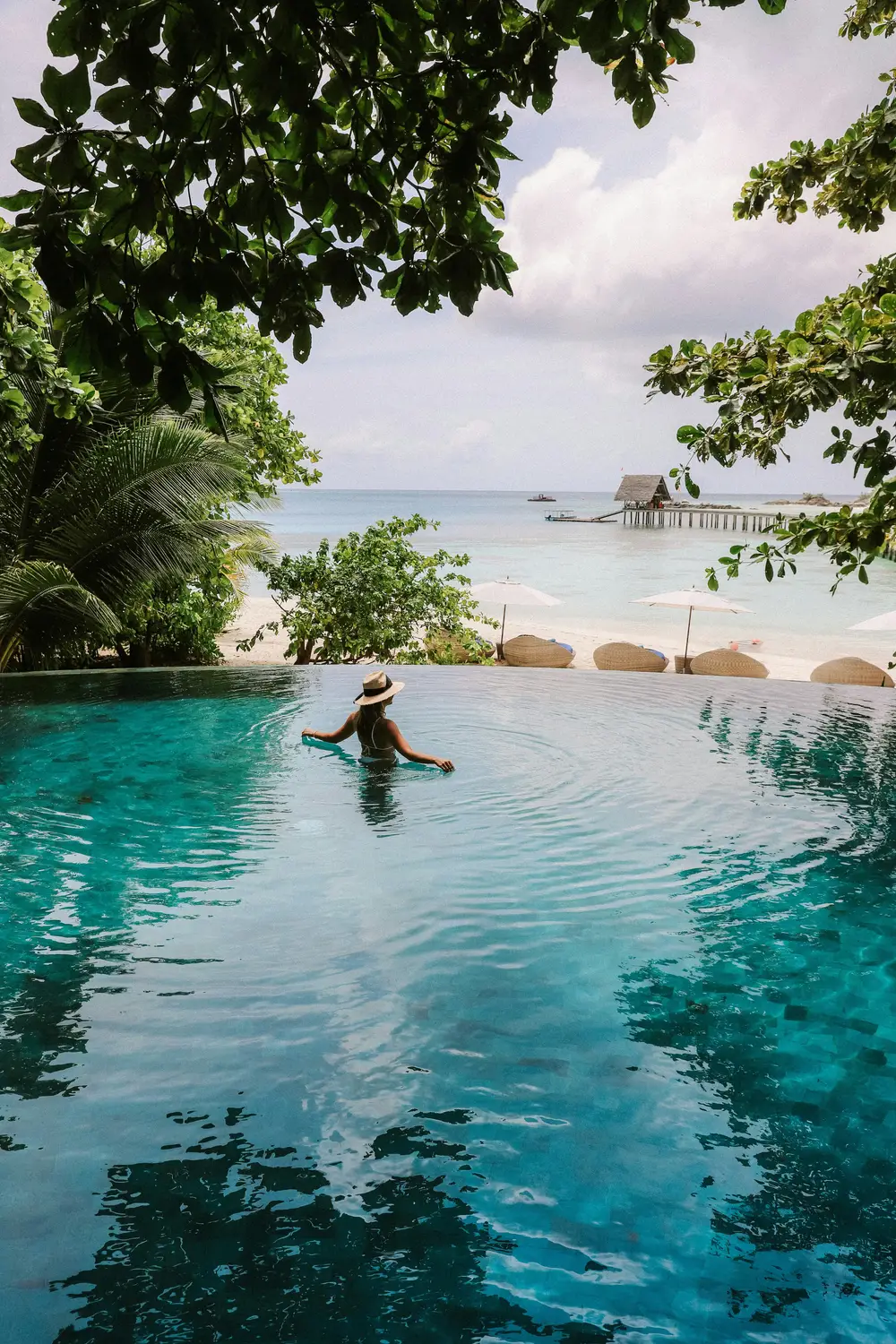 Person swimming in infinity pool overlooking turquoise ocean lagoon surrounded by tropical trees at Maldives luxury resort