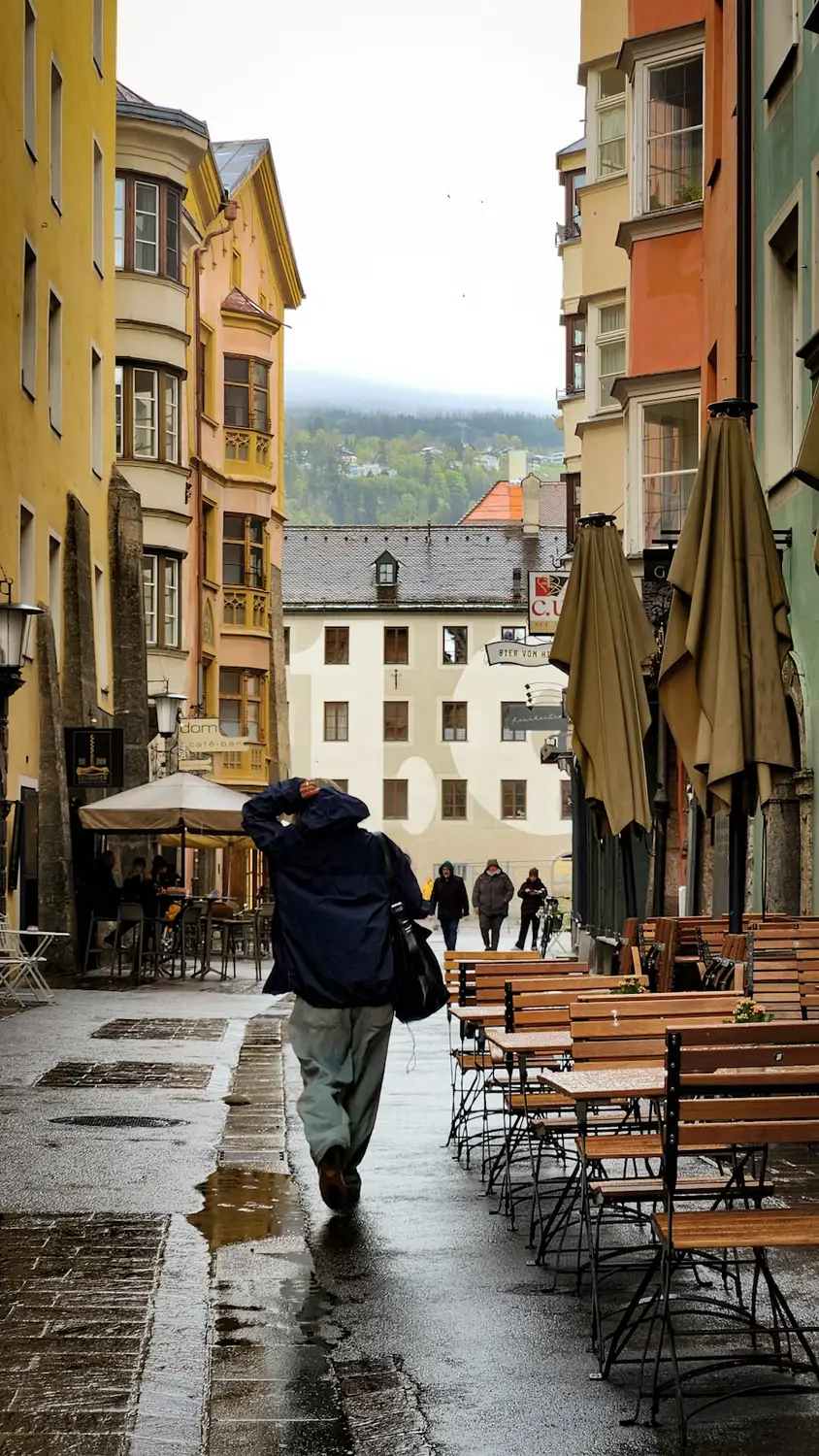 A person walking down a narrow, rain-slicked cobblestone street lined with colorful historic buildings and empty outdoor café tables.