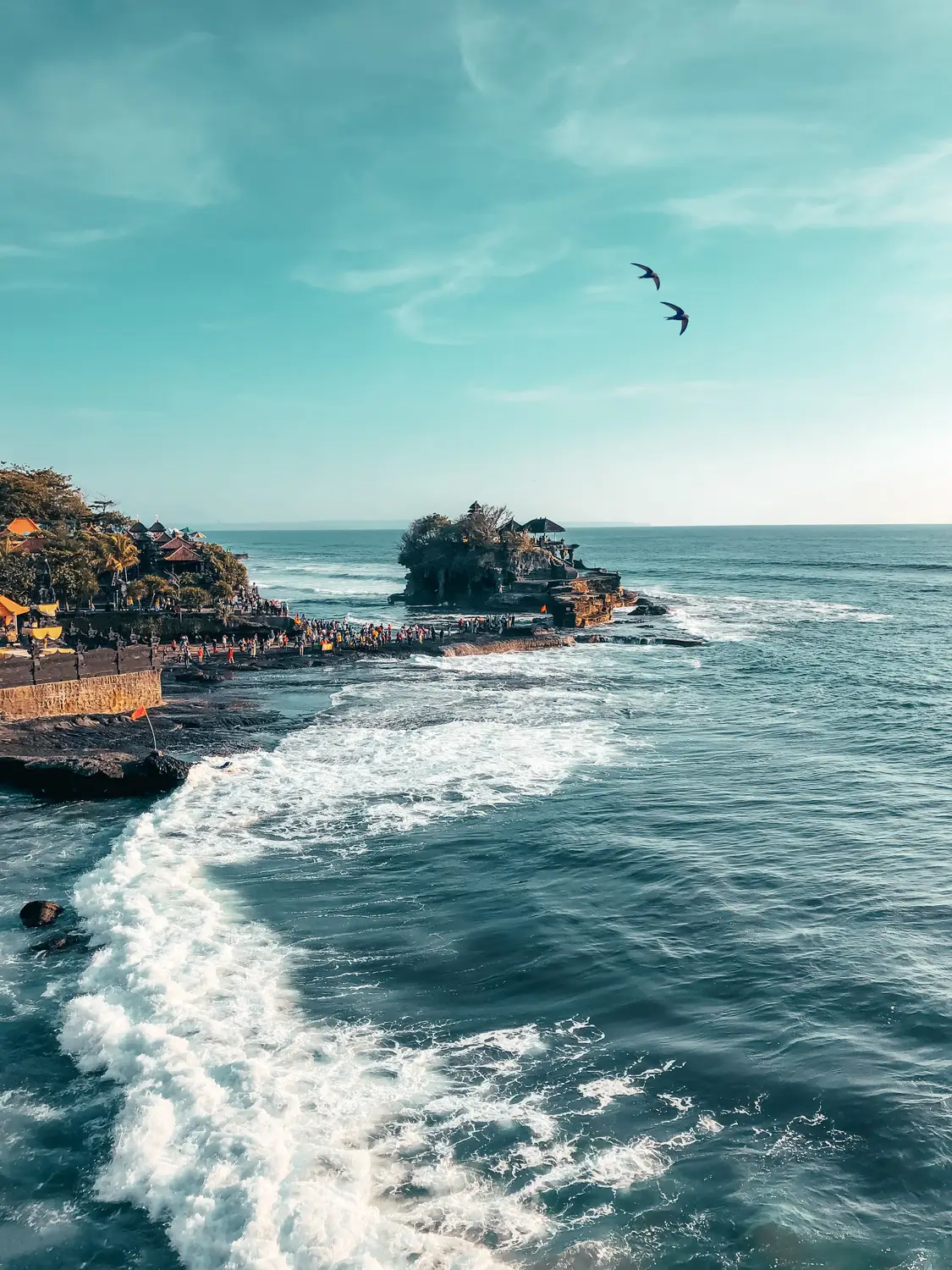 Aerial view of Tanah Lot temple on a rocky sea outcrop with crashing waves along the coastline in Bali, Indonesia