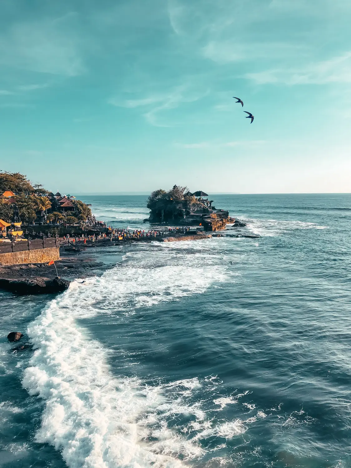 Aerial view of Tanah Lot temple on a rocky sea outcrop with crashing waves along the coastline in Bali, Indonesia