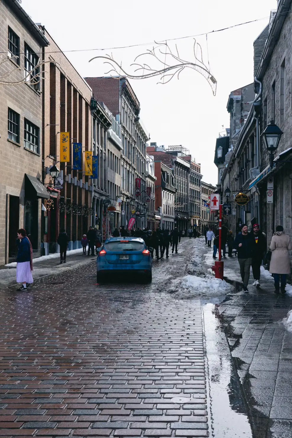 A wet cobblestone street lined with historic buildings and shops, with a few pedestrians walking in the old town.