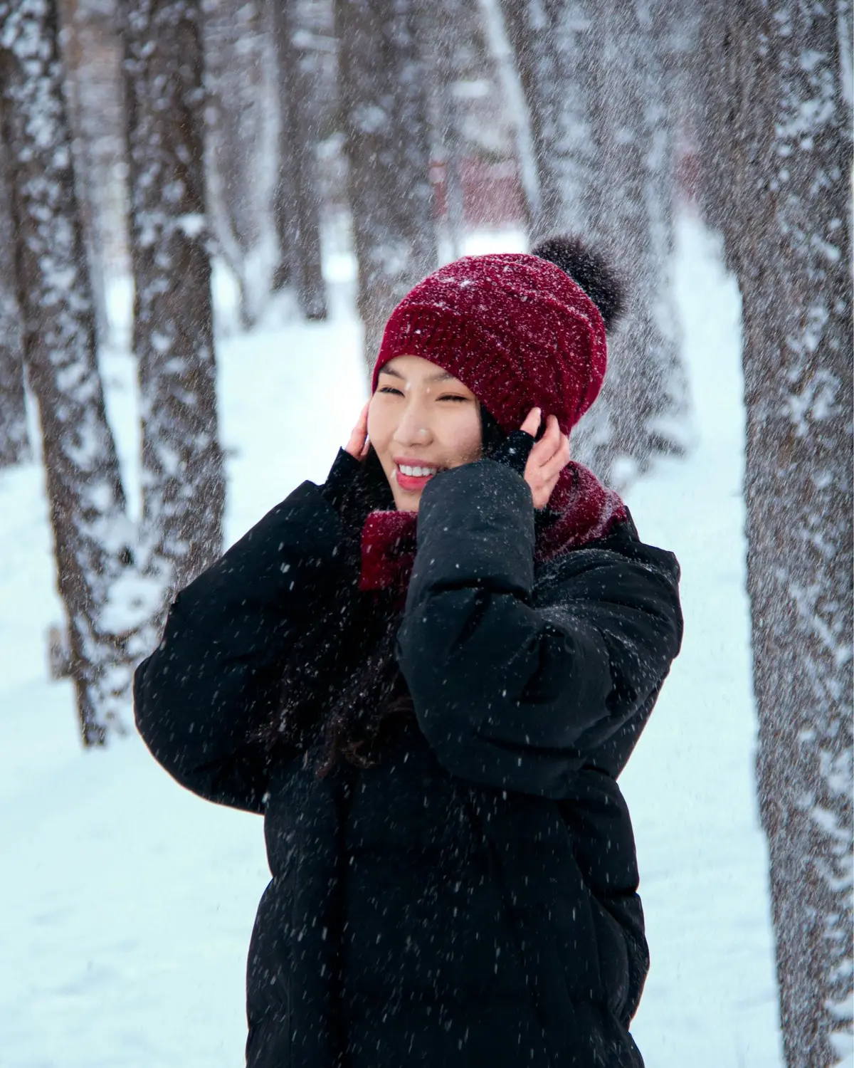 A smiling woman in a red pom-pom beanie and black puffer jacket standing joyfully in a snow-dusted winter forest with flakes falling around her