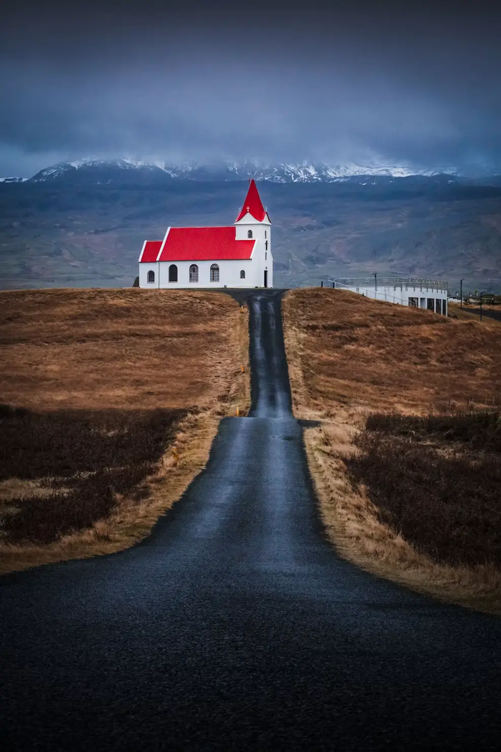 Red-roofed church at the end of a winding road through dark volcanic hills, under a stormy sky, Iceland.
