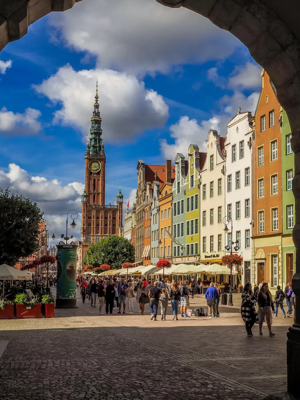 Colorful European city street with historic buildings, a church tower, and pedestrians under a blue sky.