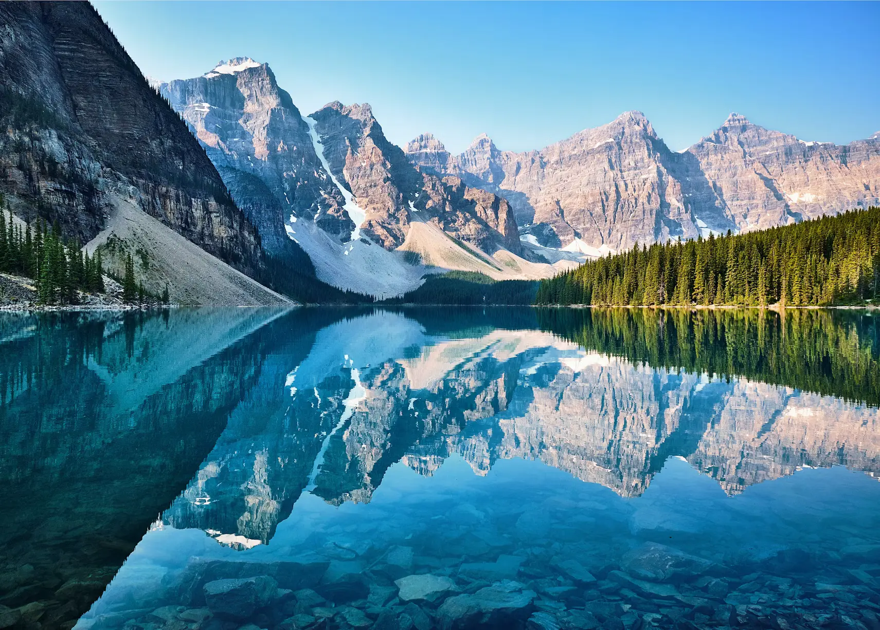 A turquoise glacial lake surrounded by snow-capped mountains and pine forest, with the peaks reflected in the still water.