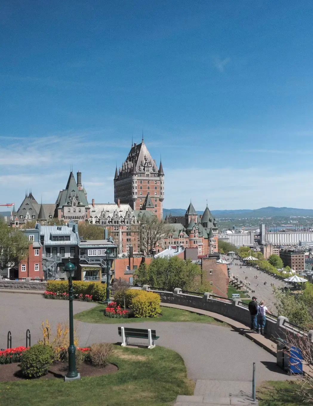 The turreted Château Frontenac hotel overlooking a riverside park with a path and benches, under a clear sky.