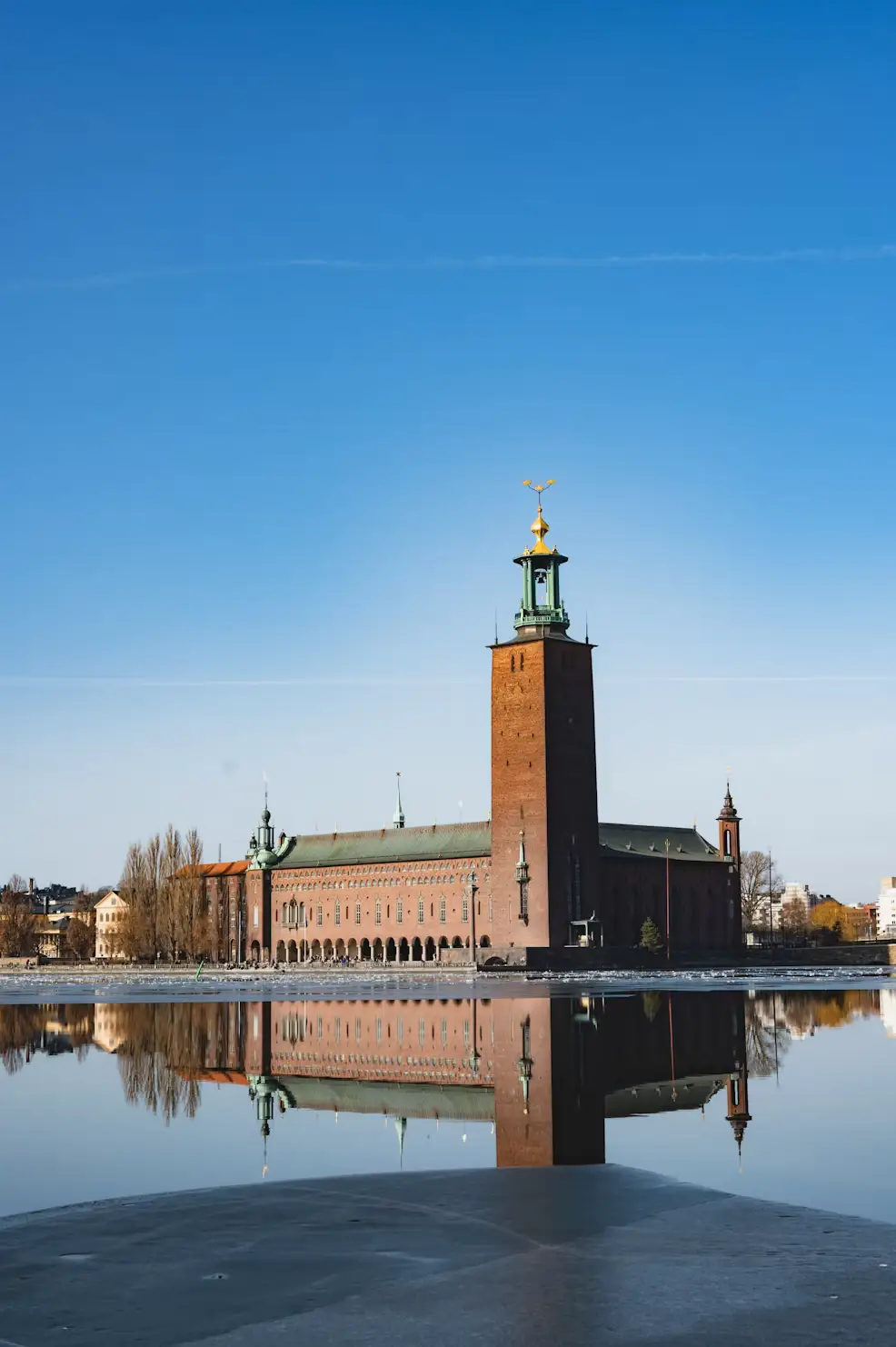 Stockholm City Hall's tower reflected in the still water beside it, under a clear blue sky.