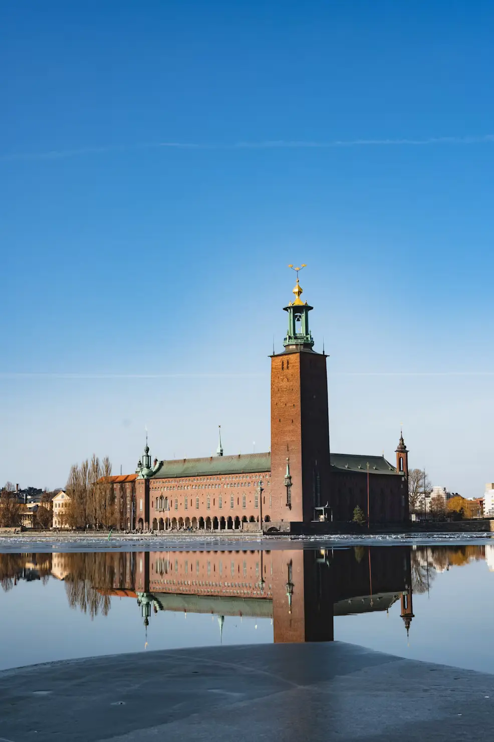 Stockholm City Hall's tower reflected in the still water beside it, under a clear blue sky.