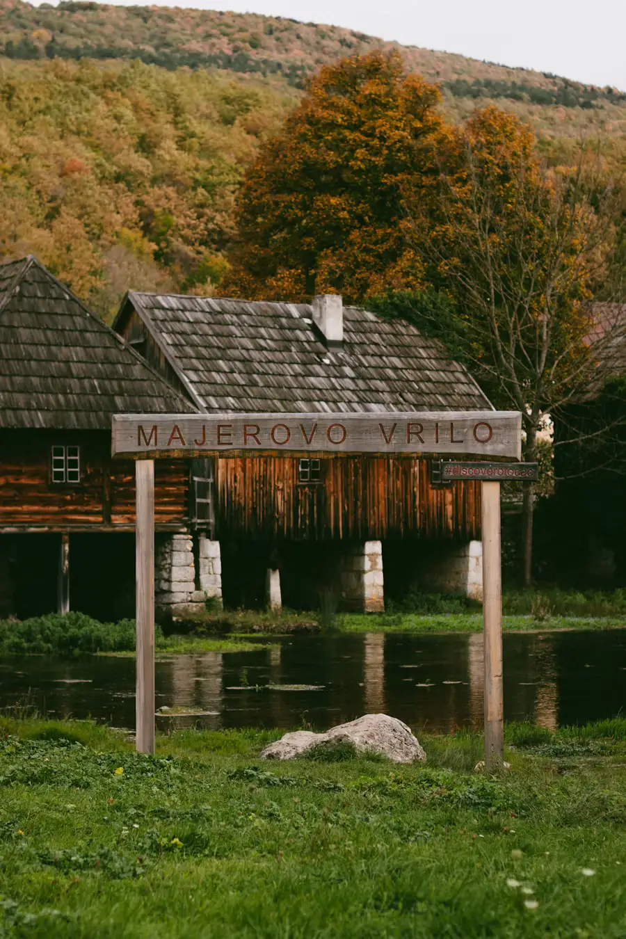 Old wooden watermill house on stilts above a stream, with an autumn-colored hillside in the background.