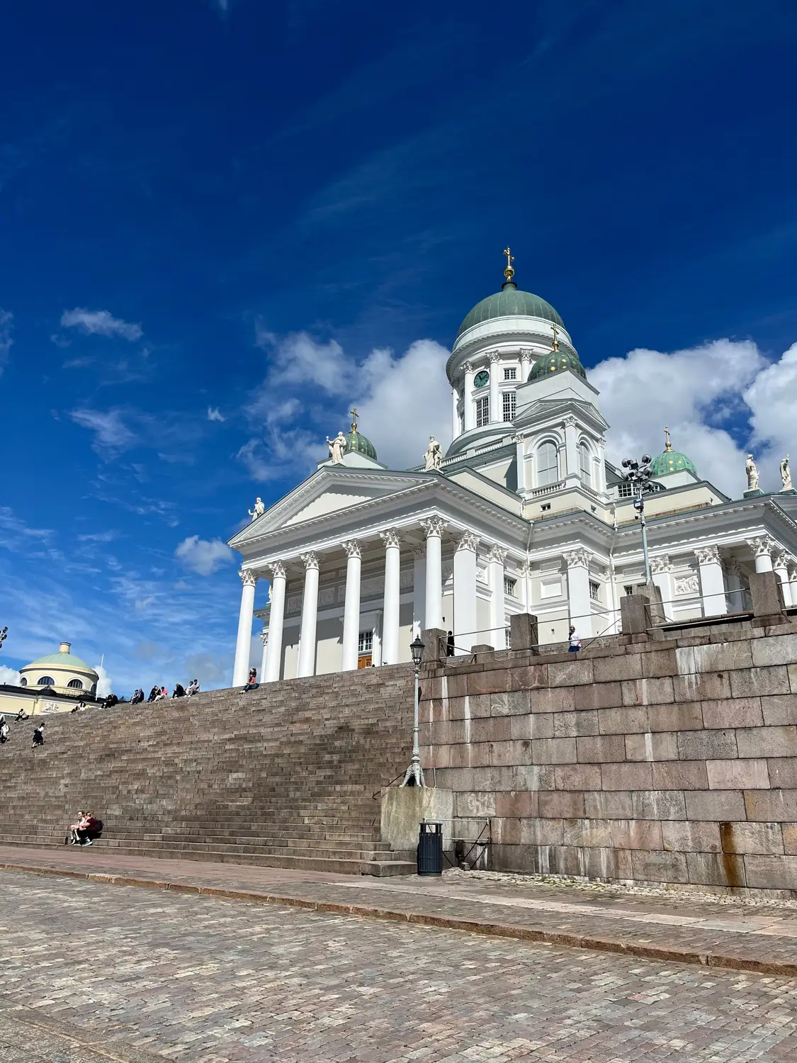 Helsinki Cathedral with its grand white columns and green domes under a vivid blue, cloud-filled sky.