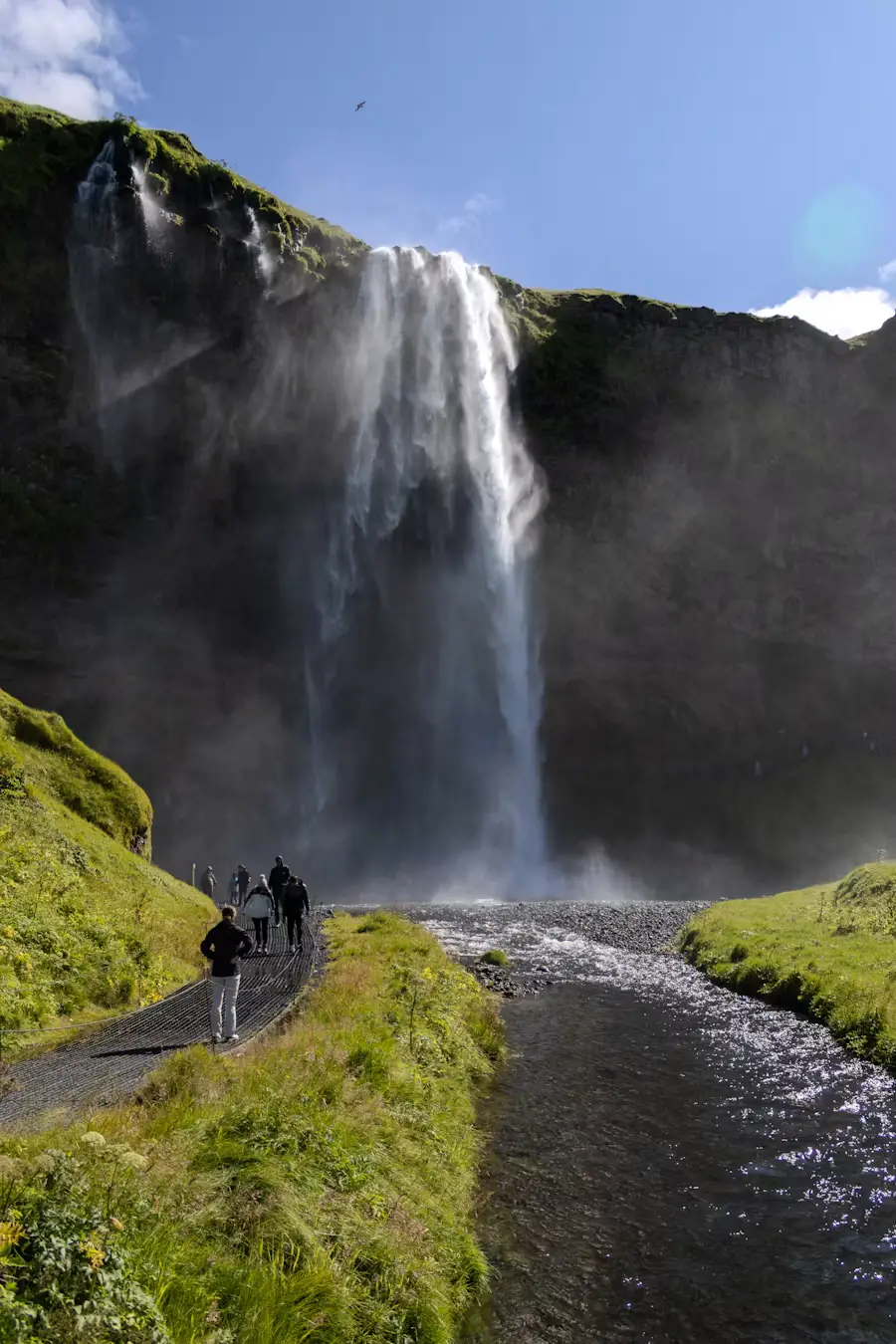 Waterfall dropping from a cliff edge into a stream, with people walking along the grassy path beside it, Iceland.
