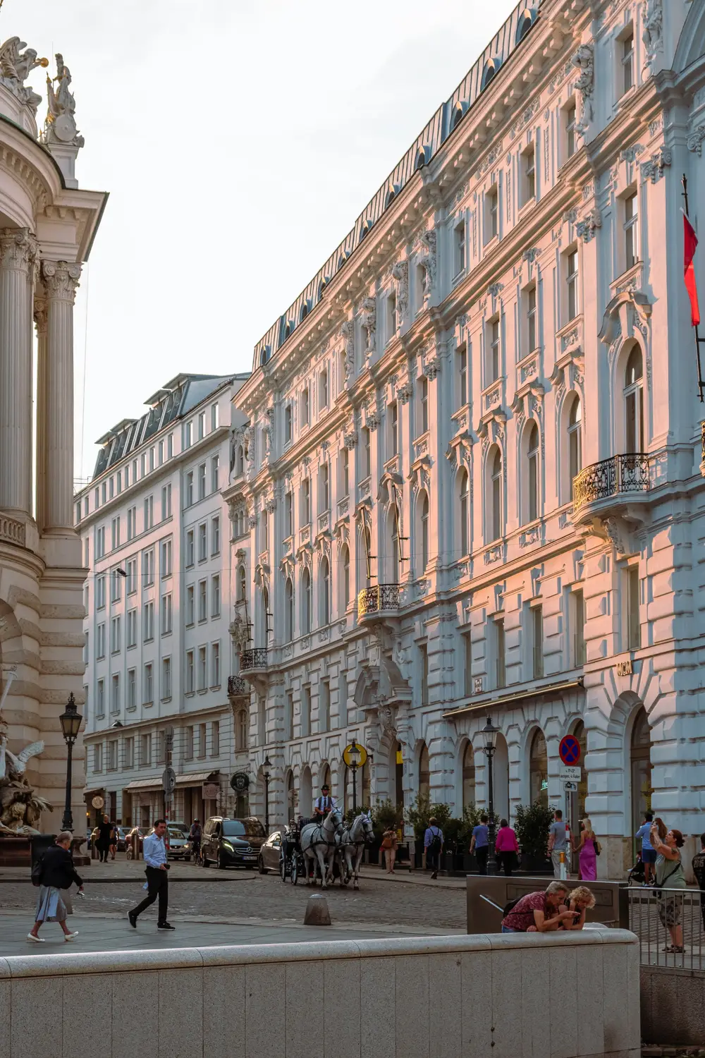 A grand European classical building facade with pedestrians walking along a paved plaza in front of it.