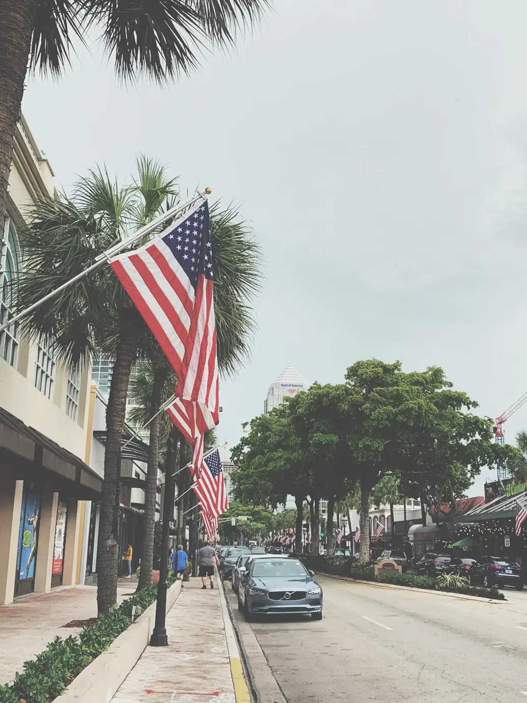 American flags hanging over a palm-lined street of storefronts on a hazy day.