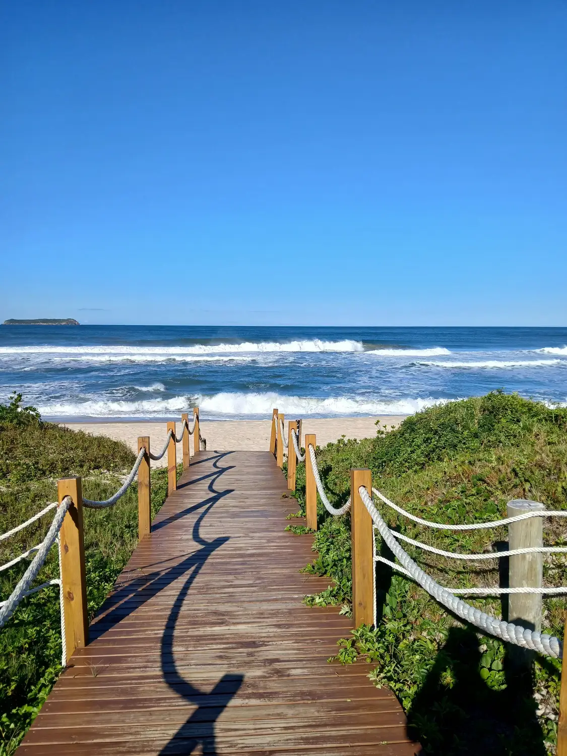 Wooden boardwalk with rope railings leading to sandy beach with rolling waves and clear blue sky