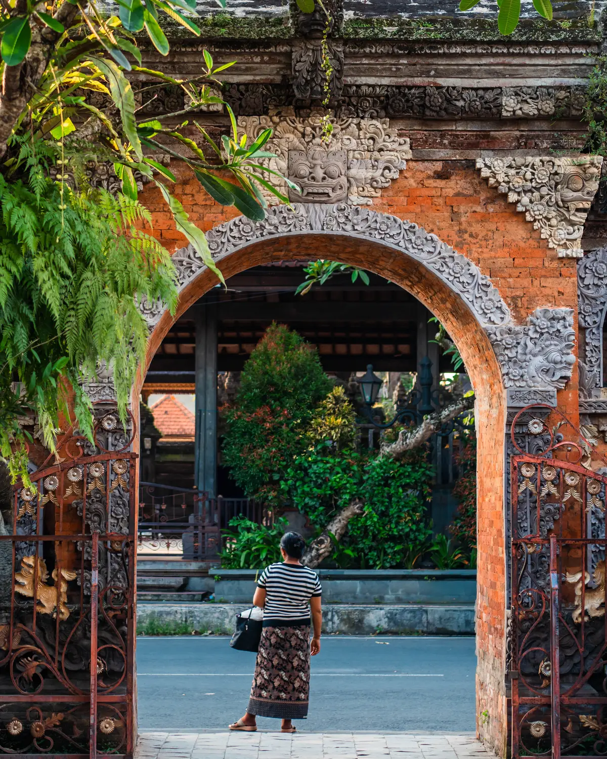 Person in traditional dress standing at an ornate red brick temple archway with intricate carvings in Bali, Indonesia.