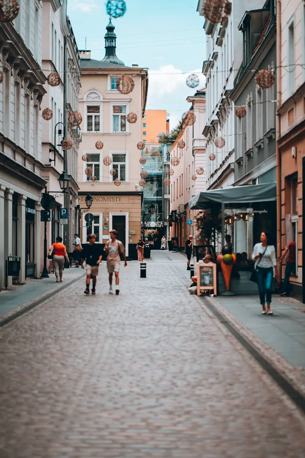 Narrow cobblestone street lined with colorful historic buildings, pedestrians strolling under a partly cloudy sky, Poland.