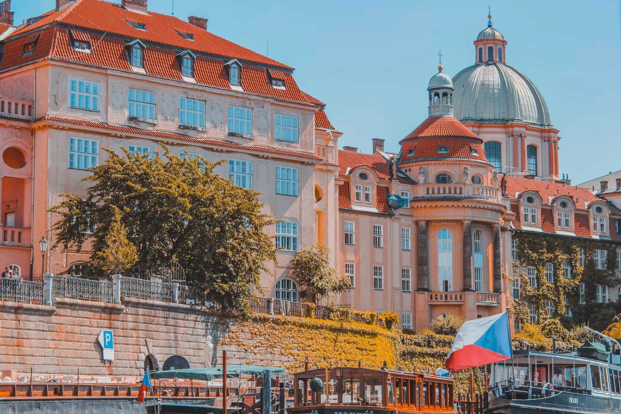 Riverside view of Prague's pink baroque buildings with red-tiled roofs along the Vltava River, with a Czech flag visible in the foreground