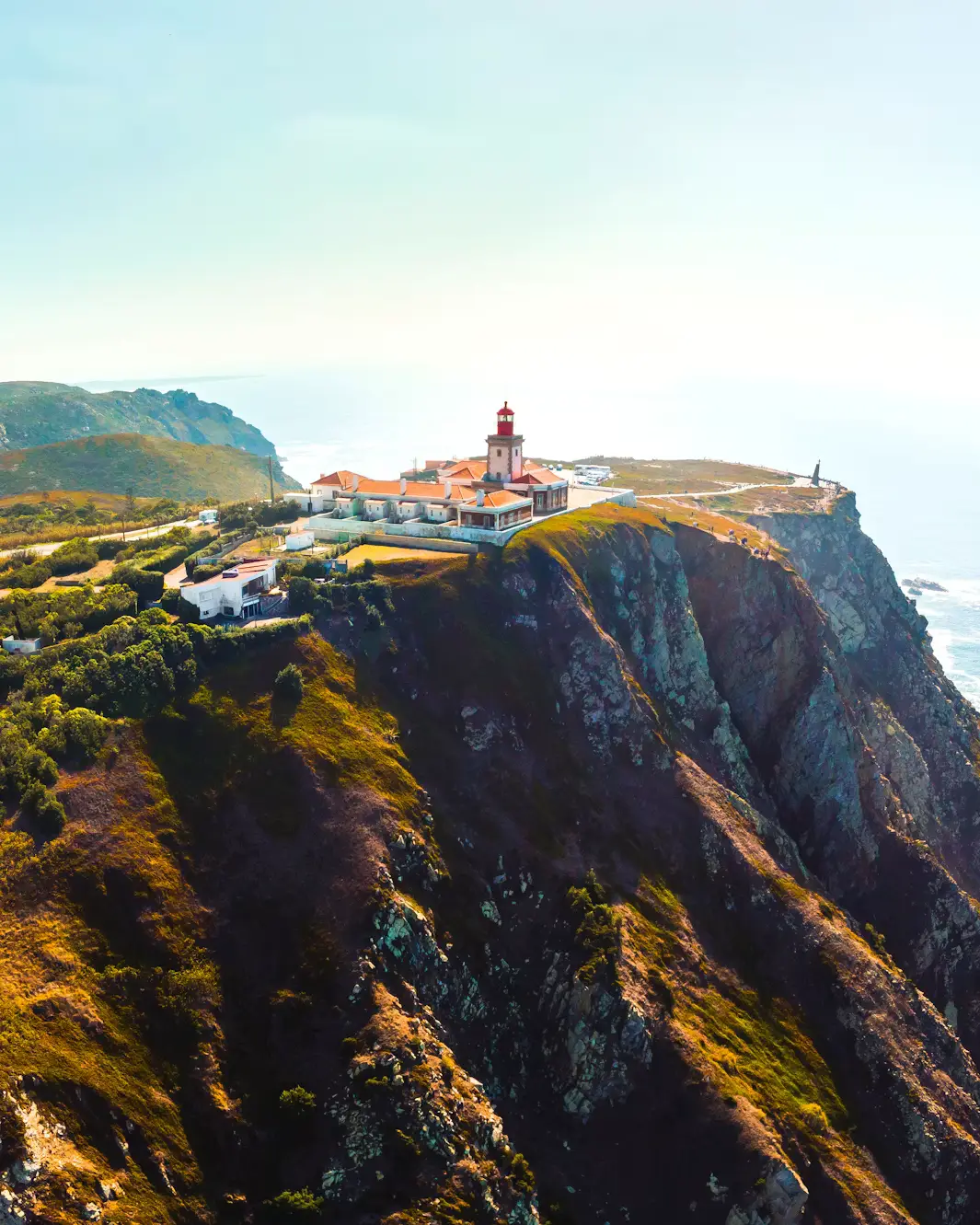 Lighthouse complex with red roofs perched on a dramatic rocky cliff overlooking the ocean.