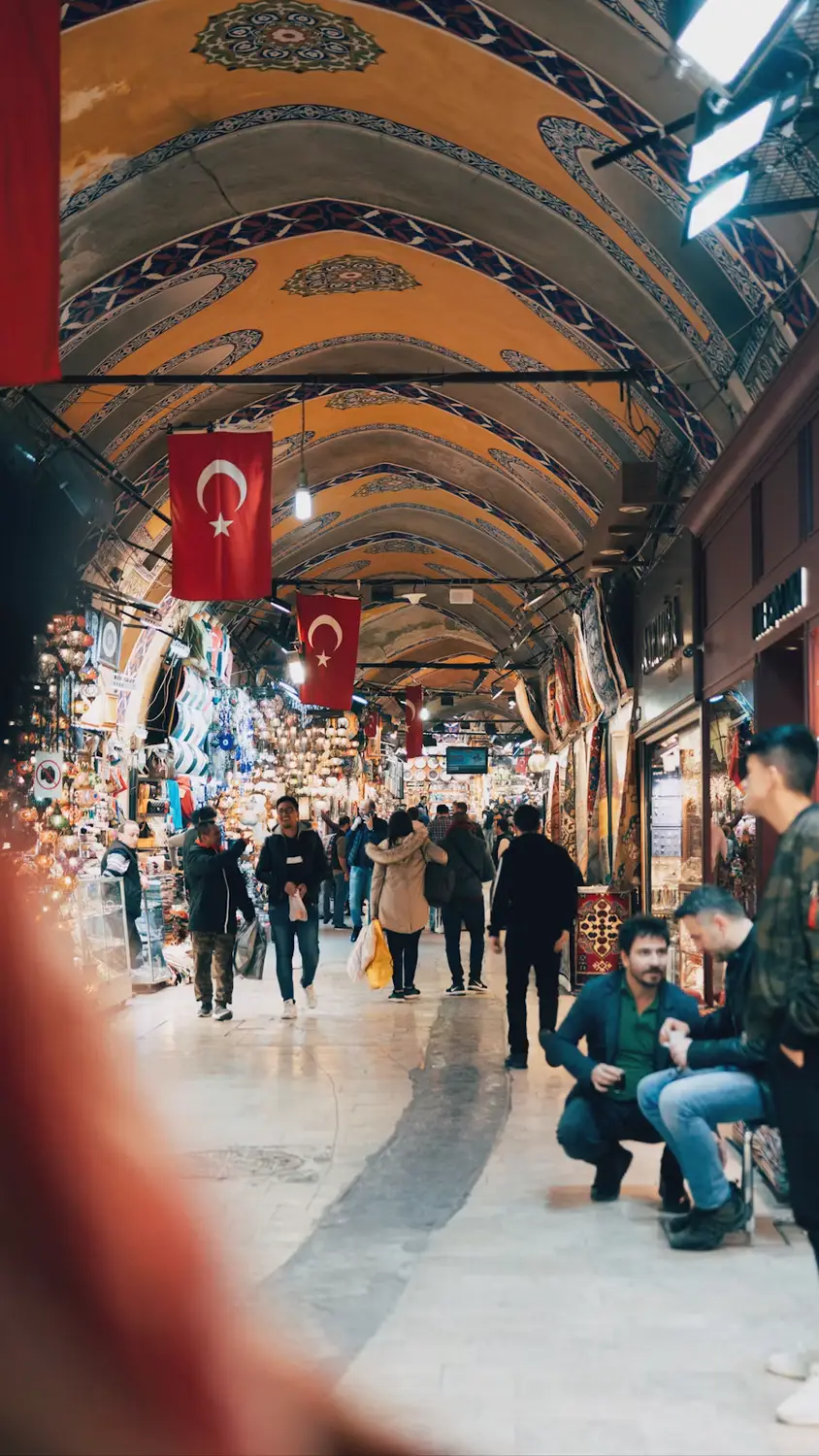 Shoppers walking through the arched, ornately painted corridor of the Grand Bazaar in Istanbul, with a Turkish flag hanging overhead.