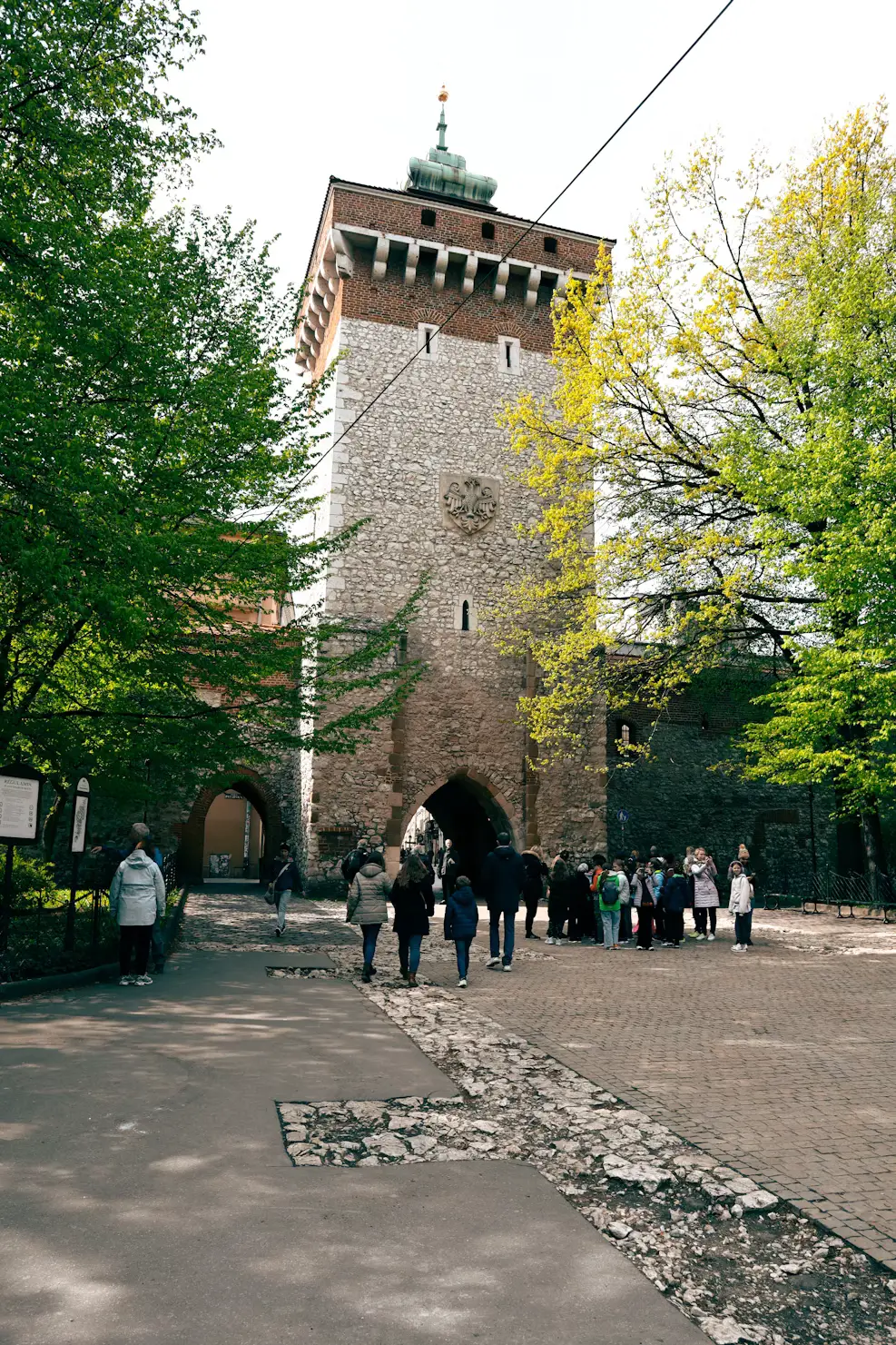 Historic stone gate tower framed by trees, with tourists gathered at its entrance, Kraków, Poland.
