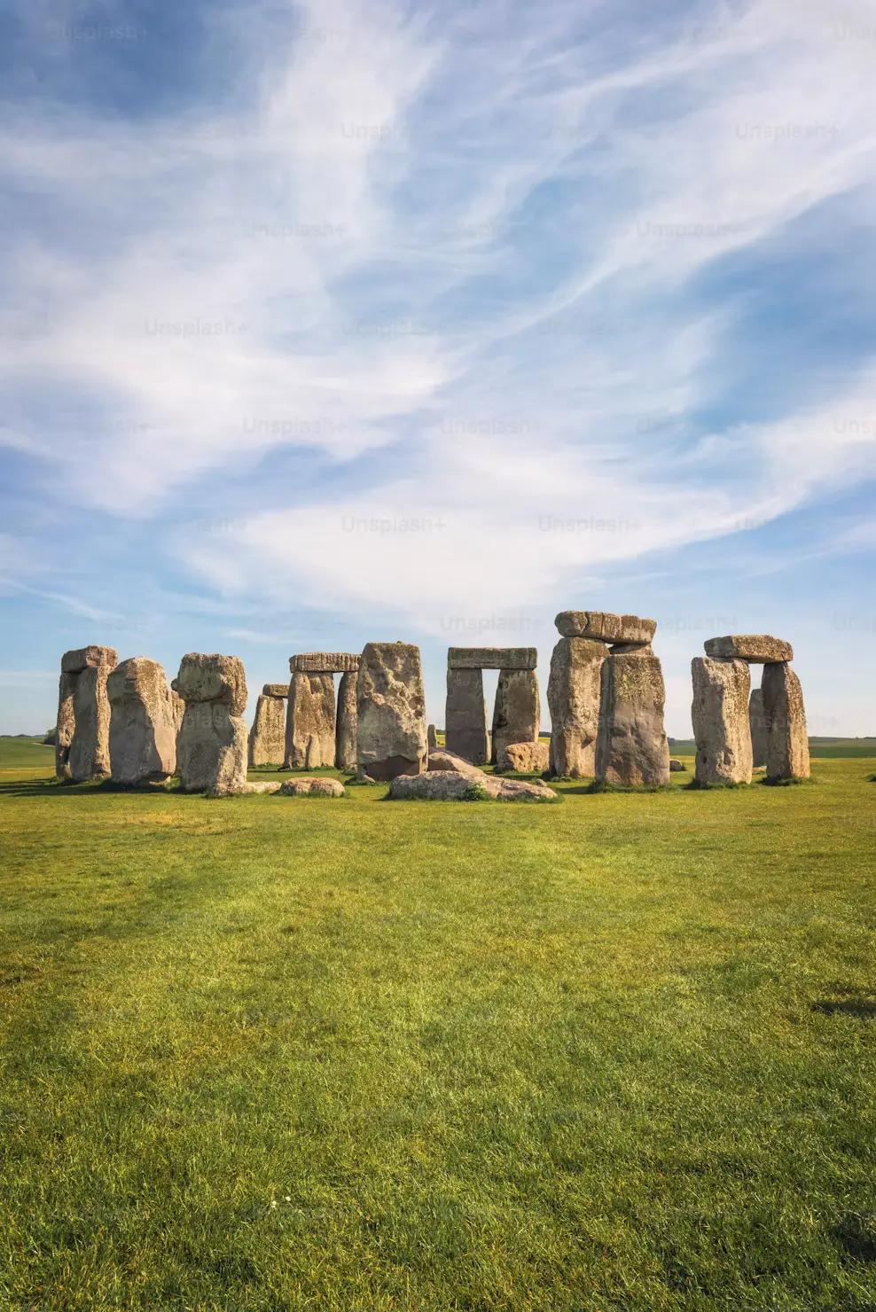 The ancient standing stones of Stonehenge on a wide green field beneath a blue sky streaked with clouds.