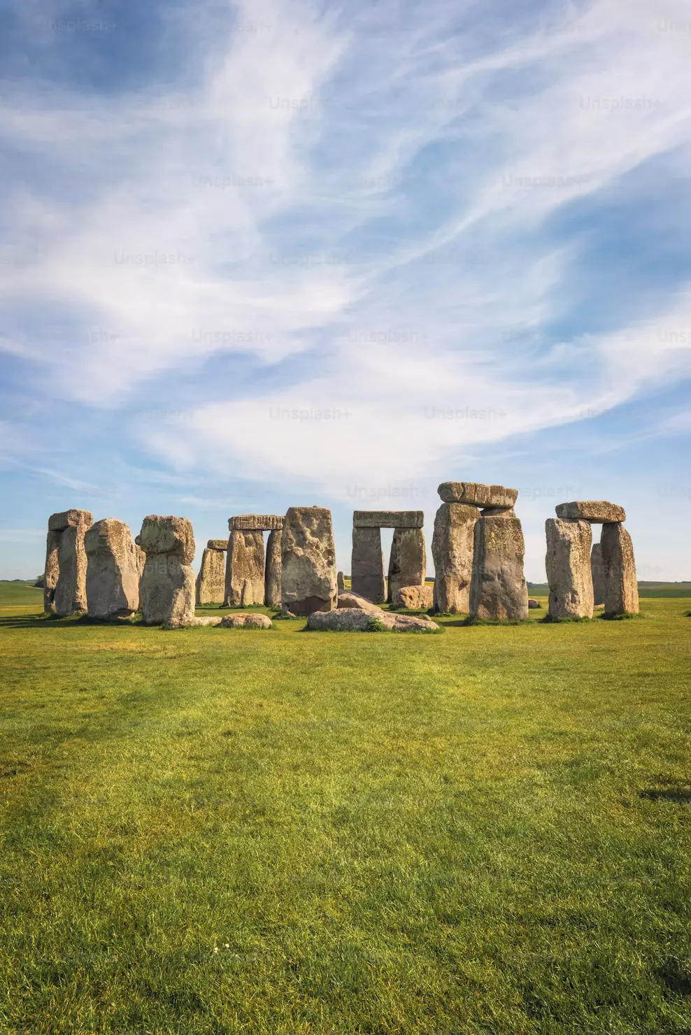 The ancient standing stones of Stonehenge on a wide green field beneath a blue sky streaked with clouds.