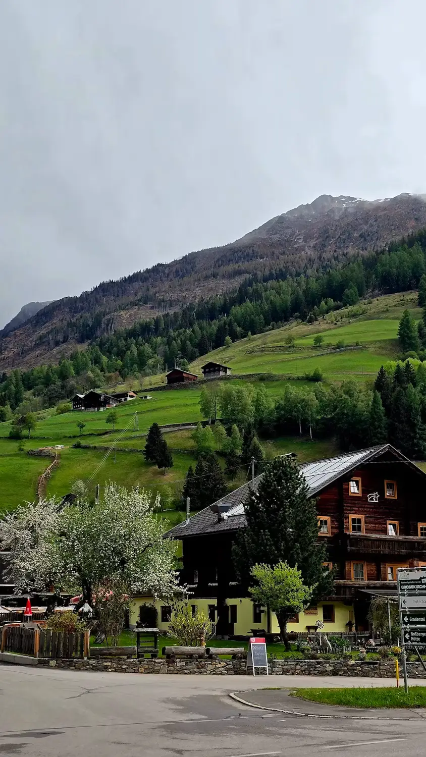 A traditional Alpine chalet with a steep gabled roof sits in a green valley, framed by grassy hillsides and mountains under an overcast sky.